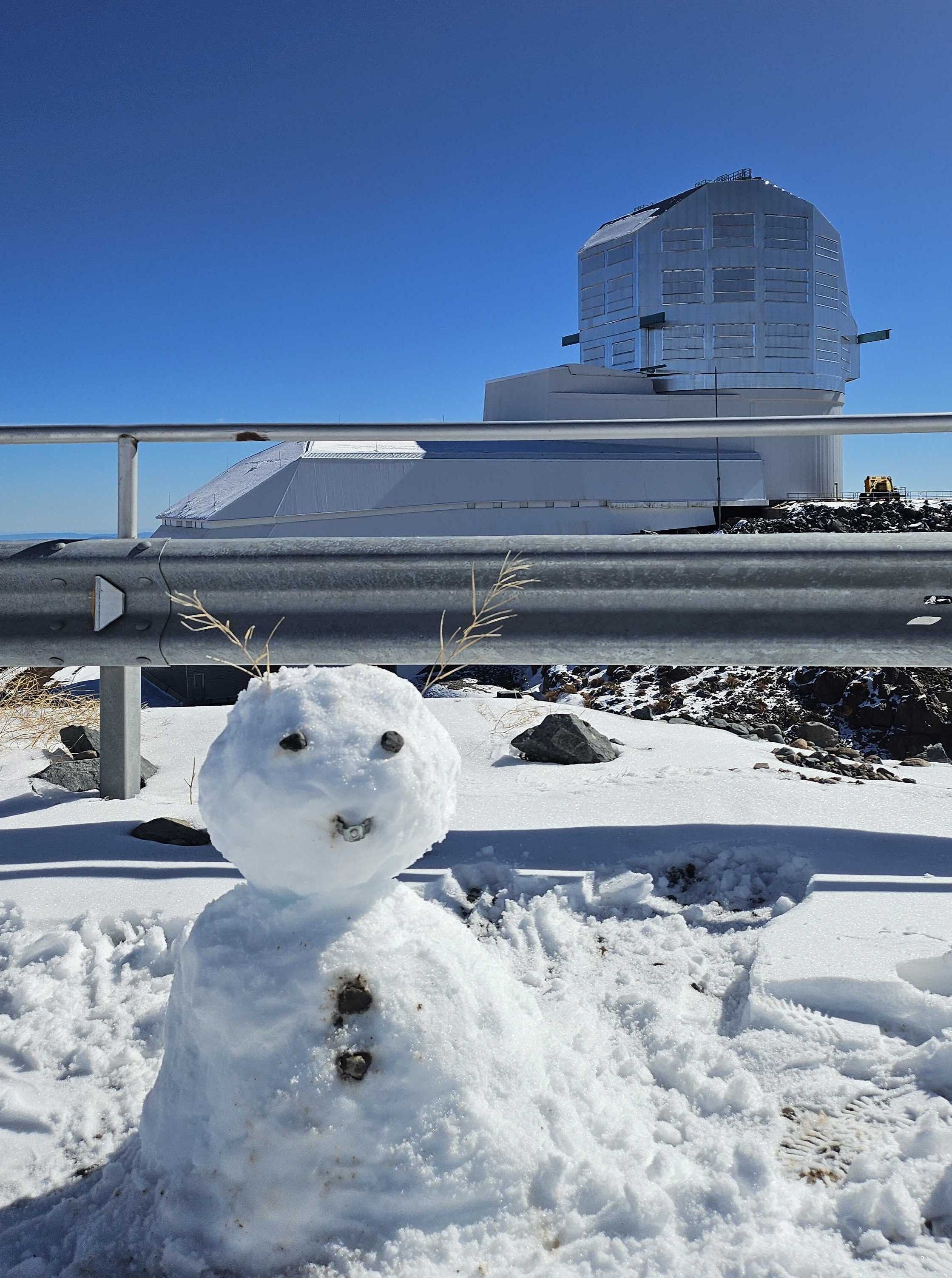 Snowman in front of Rubin Observatory. There is snow on the ground and also on the observatory dome shutter door.